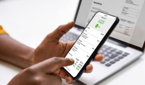 Persona usando un smartphone con una aplicación financiera frente a un portátil mostrando una factura