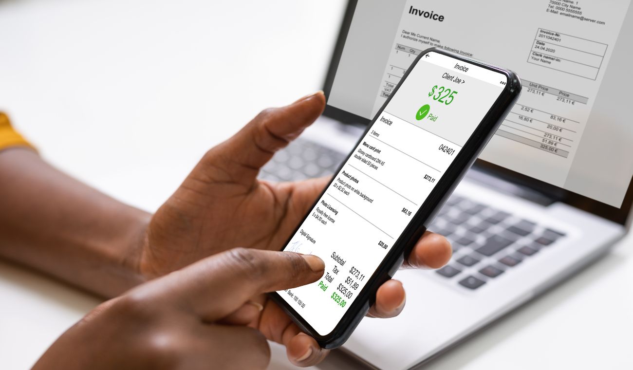 Persona usando un smartphone con una aplicación financiera frente a un portátil mostrando una factura