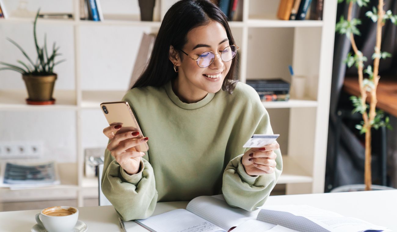 Mujer sosteniendo un móvil y una tarjeta de crédito mientras sonríe en una mesa con libros y una taza de café