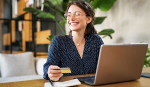 Mujer sonriendo con gafas sosteniendo una tarjeta de crédito frente a un portátil
