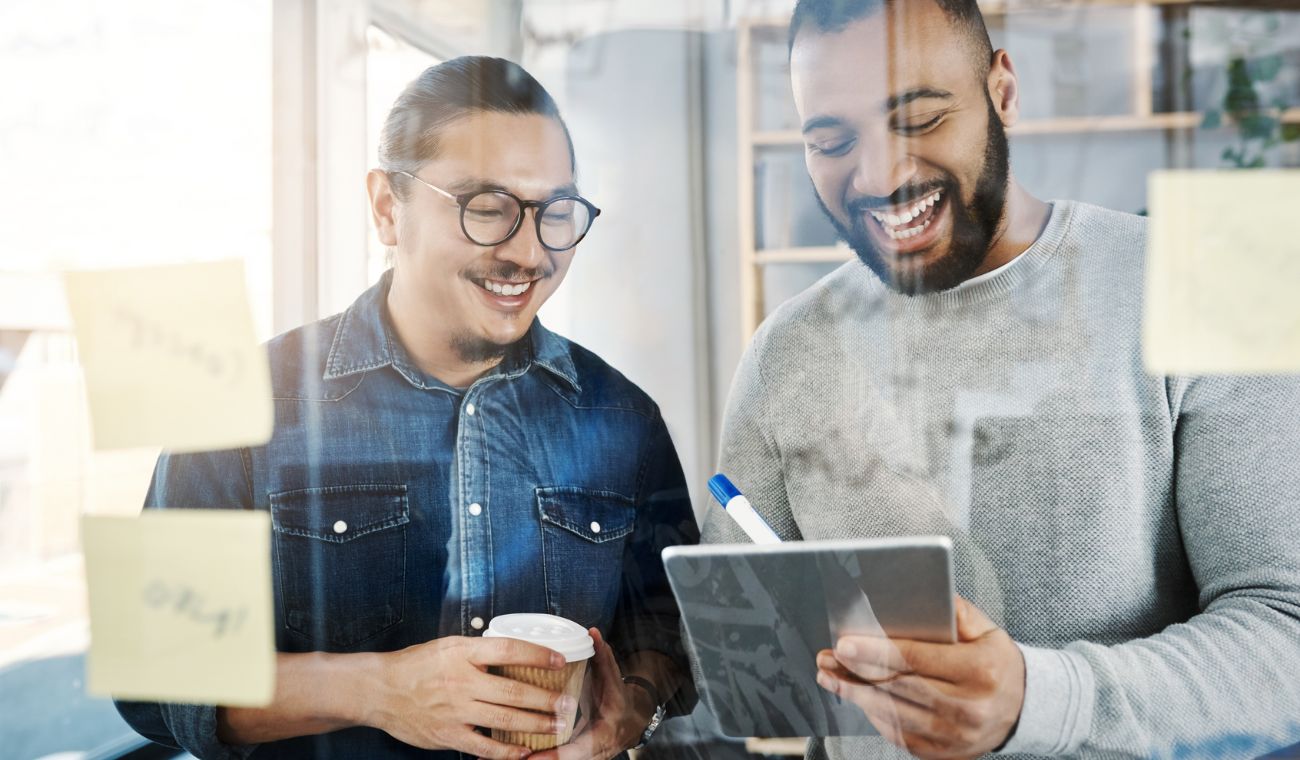 Dos personas sonriendo y revisando una tableta en una oficina con notas adhesivas en el cristal