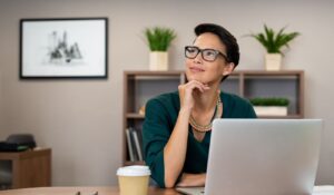 Blog 11 Persona con gafas sentada en un escritorio junto a un portátil y una taza pensando con la mano en la barbilla
