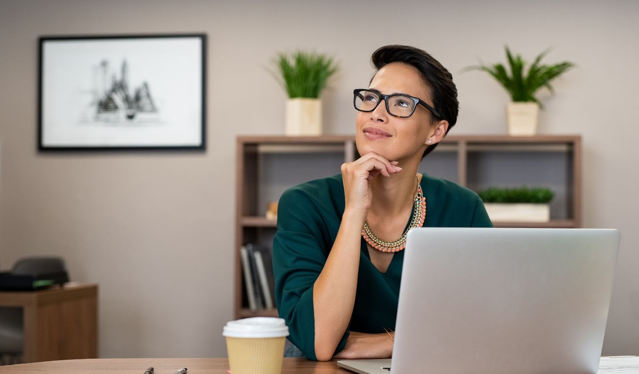 Persona con gafas sentada en un escritorio junto a un portátil y una taza pensando con la mano en la barbilla